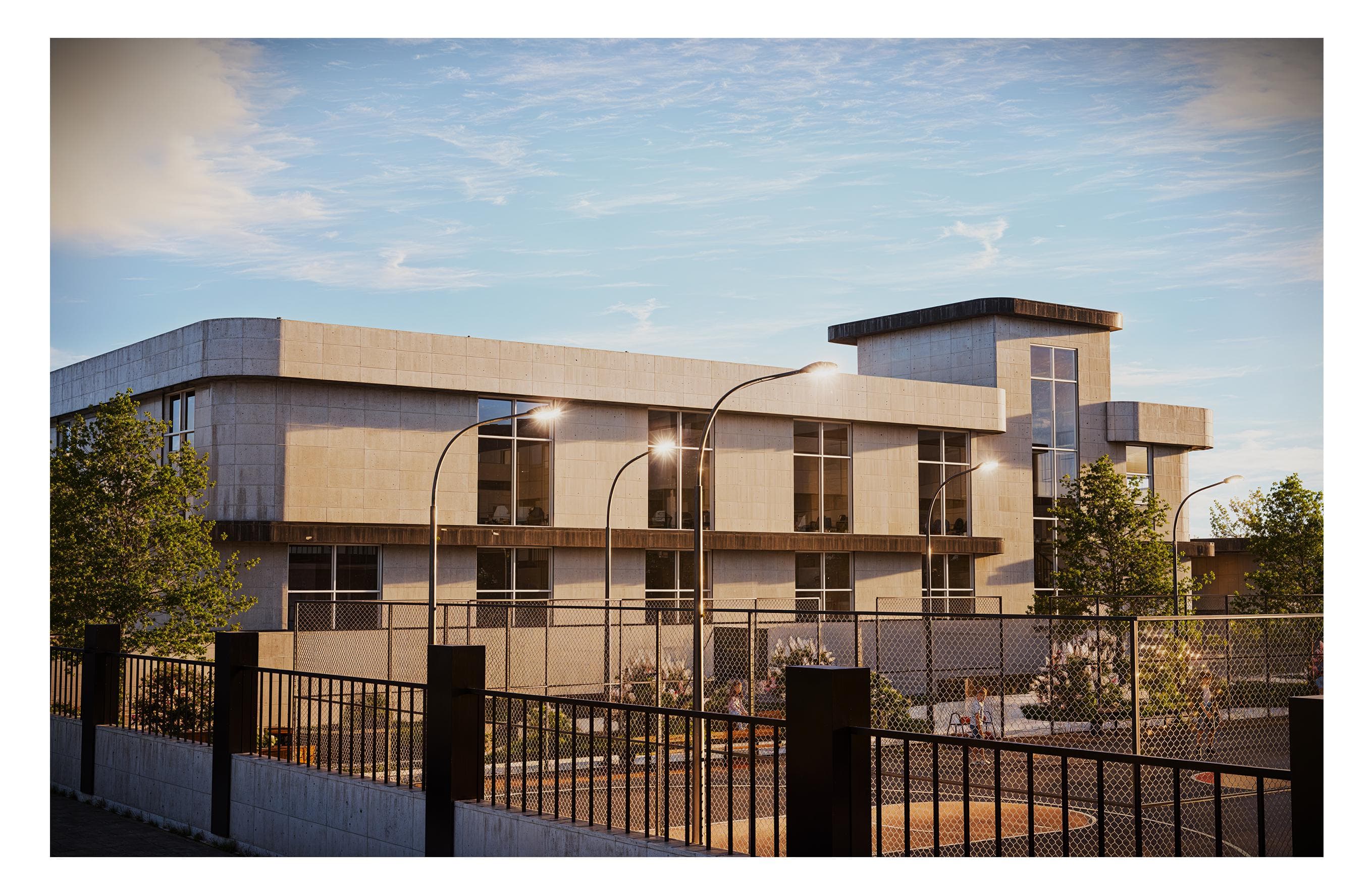 Finally, along the perimeter street, the complex presents a composed civic face—layered greenery, secure fencing, and warm evening light across the stone‑toned façades. The corner stair tower acts as a clear wayfinding marker and anchors the neighborhood edge of the campus.
